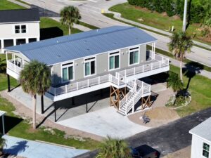 Modern double-wide manufactured home elevated on stilts with front deck and palm trees in a landscaped mobile home community.